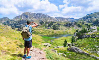 Young traveler man with a backpacker standing on an alpine mountain trail with a lake. Circo de Saboredo, Aran Valley, Catalan Pyrenees, Catalonia, Spain, Europe.