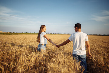 Happy couple in love walk along the wheat field in summer. A woman and a man in white T-shirts run across the field holding hands on a sunny day.