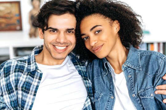 Joint photo of a hispanic guy with african american girl. Close-up portrait of happy young mixed race family couple sitting in living room, wearing casual stylish clothes, looking at camera, smiling