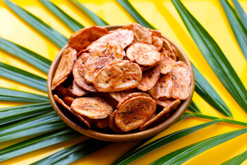 Banana chips in wooden bowl on palm leaves on yellow background.