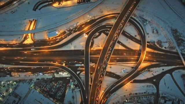 Car traffic on illuminated road junction top down aerial view. Winter night city highway. Snow town motorway with trucks lights. Urban infrastructure. Twilight cityscape. Europe cinematic drone shot