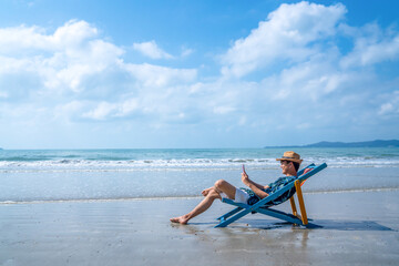 Asian man resting on sunbed on tropical beach. Happy guy sitting on beach chair by the sea using...