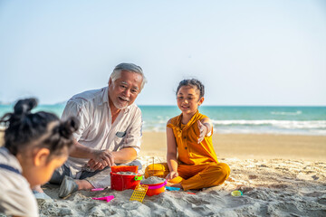 Happy Asian family on summer travel vacation. Grandfather enjoy play beach toys with two grandchild girl on sand beach in sunny day. Senior man with granddaughter having fun outdoor lifestyle activity