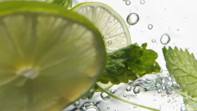 Slices of fresh lime and mint leaves fall down into water with bubbles on white background macro bottom view slow motion