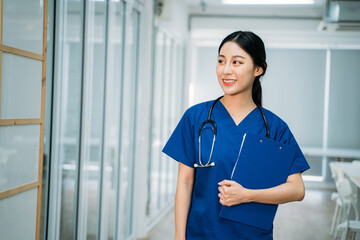 Young beautiful Asian female doctor in uniform with stethoscope standing in hospital hallway holding clipboard checking reports while looking away