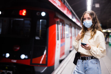 Obraz na płótnie Canvas Portrait of a thoughtful girl in a protective mask, standing on the platform of a subway station during a pandemic ..with a mobile phone in her hands while waiting for a train