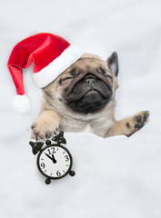 Pug puppy wearing red santa hat sleeps with alarm clock under white blanket at home. Top down view