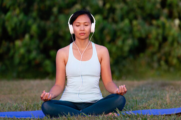 people, fitness, music and healthy lifestyle concept - asian girl meditating and listening the music with headset in the park