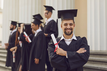 Happy confident man in graduation gowns holding diploma and smiling while his friends standing in the background. Graduation from the university. Education, university and college concept. © Studio Romantic
