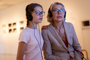Senior female tutor and tweenage girl exploring art pieces in museum and listening commentaries in headphones
