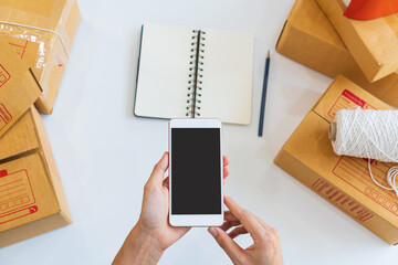 Obraz premium Hands of woman entrepreneur holding smartphone with cardboard boxes, computer laptop, online selling equipment. Preparing for shipping, Packing, business and technology concept. Top view, copy space