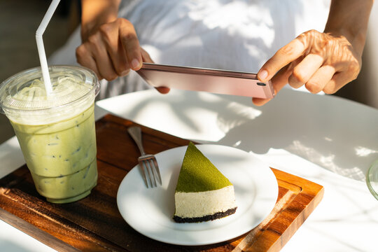 Woman hands using smartphone to take a picture of Matcha cheese cake with iced Matcha latte on white table in sunny day.