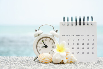 white analog clock and opened calendar with blurred shell and plumeria flower on blurred sea...