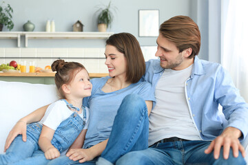 Positive friendly young parents with smiling little daughter sitting on sofa together while relaxing at home on weekend.