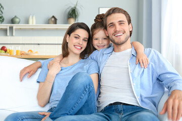 Positive friendly young parents with smiling little daughter sitting on sofa together while...