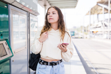 Positive european girl in blue jeans standing on station, waiting for arriving tram