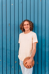 vertical portrait of a young caucasian man looking at camera smiling and holding a basketball. He has long red hair and wears casual summer clothes. It is leaning on a blue metal door