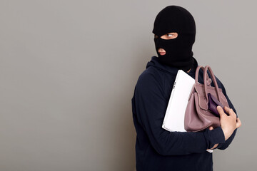 A young thief male dressed in a black hoodie with a disguised face runs away after stealing with other people's belongings, bag, laptop, wallet. Look around, be afraid of being caught, copy-space.