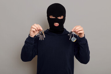 A criminal man in a black hoodie and a mask holds a bunch of keys in both hands, is about to open...