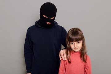 Terrible criminal man wearing black hoodie and robber mask holds a little preschool girl hostage,...