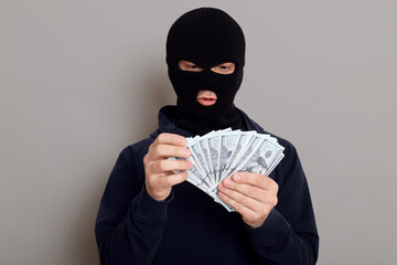 Robber man dressed in black hoodie stands with disguised face and holds a lot of money in his...