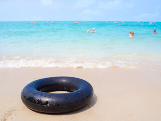 Obraz na płótnie Canvas Black swim ring or inflatable rubber toy on sand beach at coast with blured people swimming in blue sea. accessories for tourist vacation summer travel in holidays.