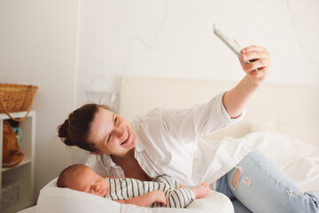 Obraz premium Cute caucasian mom with a newborn baby. A mother in a white shirt takes a selfie on the phone, the baby lies in a cocoon in a cradle for newborns , light toning
