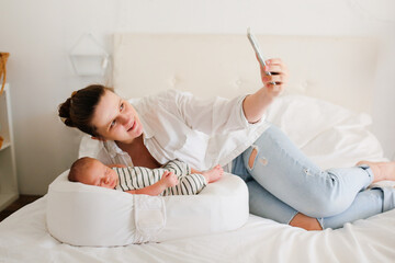 Cute caucasian mom with a newborn baby. A mother in a white shirt takes a selfie on the phone, the...