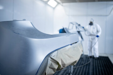 Car painter in protective clothes and mask painting automobile bumper with metallic paint and...