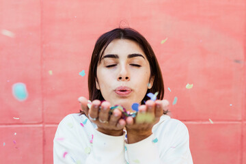 Beautiful hispanic young woman blowing confetti with pink background - Celebration, party and...
