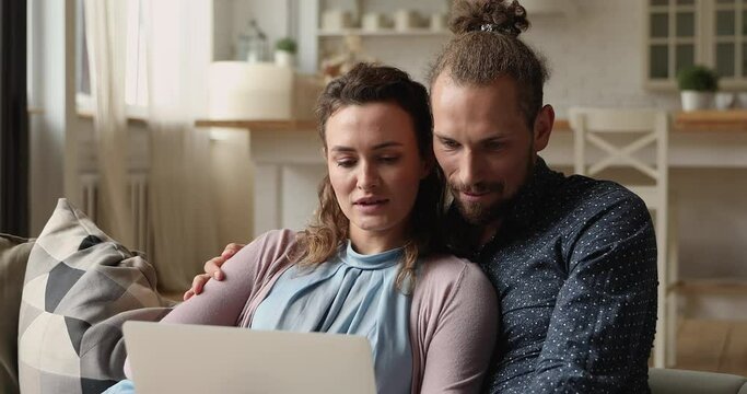 Happy affectionate bonding young married couple using computer, relaxing on comfortable sofa, planning trip vacation holidays, booking tickets, watching funny photo video content in social network.