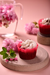Red velvet muffin with cream on a gray plate, next to sakura flowers, on a powdery background, in the background a jug with water, flowers and a muffin.