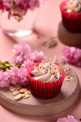 Cupcake Red velvet with almond petals on a gray plate, next to sakura flowers and almond petals, on a pink background, another cupcake in the background, side view.