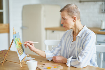 Creative hobby. Side view of concentrated mature woman painting picture at home while sitting at wooden table in kitchen, selective focus on female. Senior people and art concept