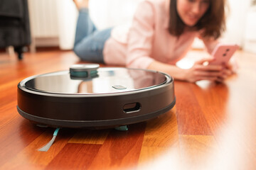 Faceless middle section of young woman using automatic vacuum cleaner to clean the floor, controlling smart machine housework robot with smart phone at home