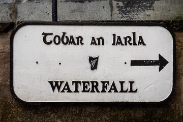 Sign waterfall in Irish and English language and arrow pointing to the right , harp emblem in the middle. White surface and black letters. Mounted on a stone and brick wall