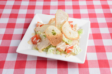 Caesar salad with chicken and greens on wooden table with red plaid table cloth.