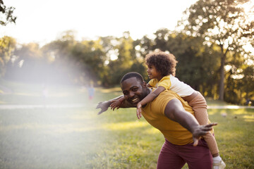 African American father and daughter having fun outdoors. Father carrying daughter on piggyback.