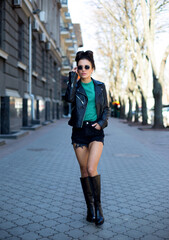 female in black leather jacket, shorts, high boot and fashionable sunglasses is walking along the...