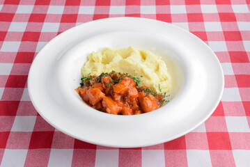 Mashed potato with gravy poured over. Served in a white plate over red plaid table cloth.