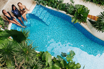 Cheerful attractive young women relaxing on edge of big hotel swimming pool and looking up at camera