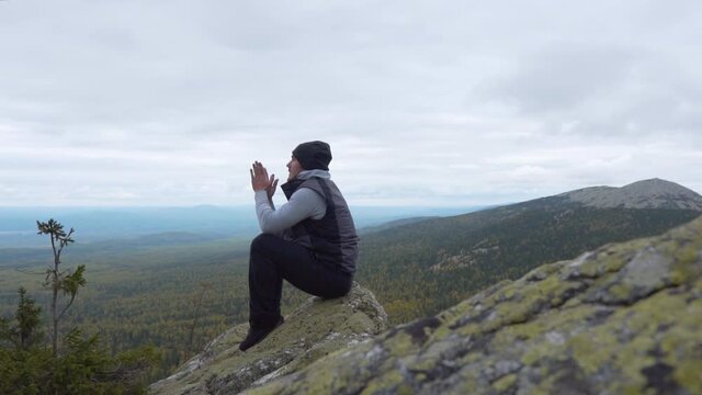 A Man At The Top Of A Cliff, Meditates With Clasped Hands Together And Relaxes And Gains Calm. The concept of Solitude and Love for Nature. Beautiful Mountain Landscape
