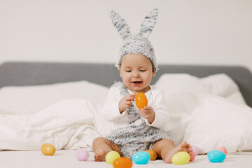 Baby sits on a bed in a bunny costume and plays with colorful easter eggs, laughs and has fun