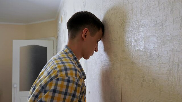Depressed guy in checkered shirt hits room wall with head tears wallpapers and beats fists with despair against white door close view