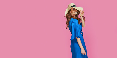Stylish fashion portrait of trendy casual young woman in blue summer dress and hat, posing over pink background.