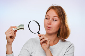 A thoughtful woman looks through a magnifying glass at a small wad of dollars. Lilac background. The concept of finance