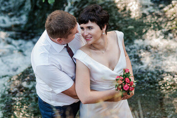 The bride and groom stand against the background of a waterfall. Newlyweds on the background of a waterfall