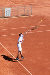 Young tennis player jump and hit ball while serving on orange clay court at start of match. Athlete in motion during game. Vertical sports background, banner, copy space