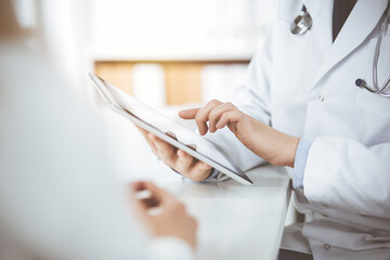 Unknown male doctor and patient woman discussing current health examination while sitting in sunny clinic and using tablet computer. Perfect medical service in hospital