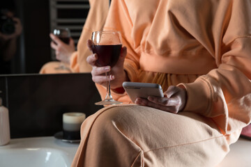 Attractive young woman in a bathroom communicating on a smartphone and drinking wine. Relaxation and leisure concept.